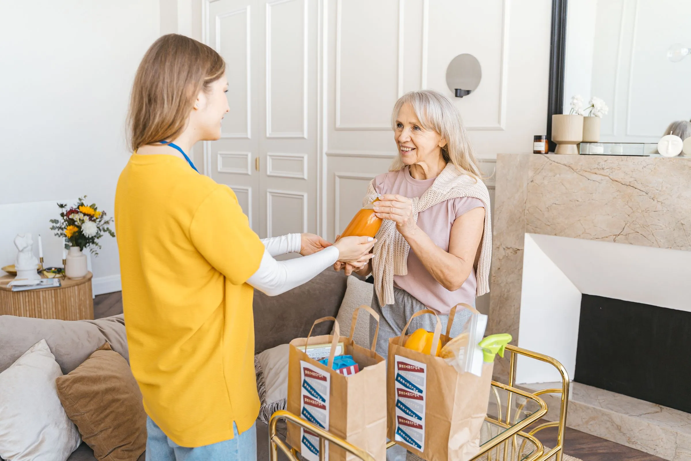A caregiver in a yellow sweater hands a bag of groceries to a smiling older woman in a bright living room