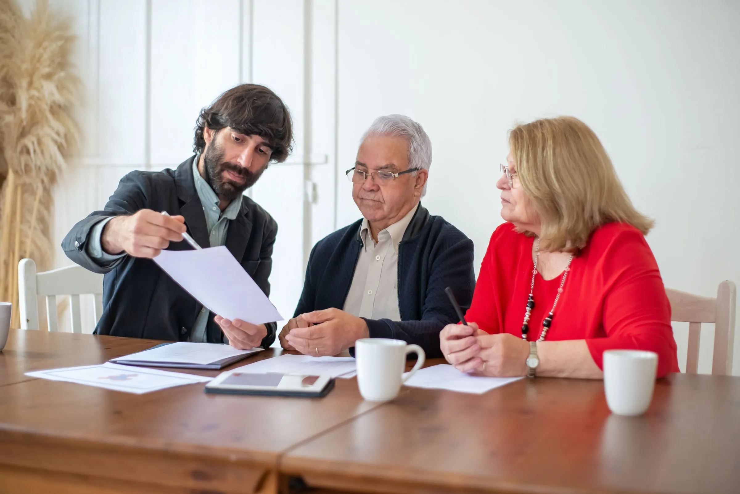 An advisor hands a sheet of paper to an older couple at a wooden kitchen table