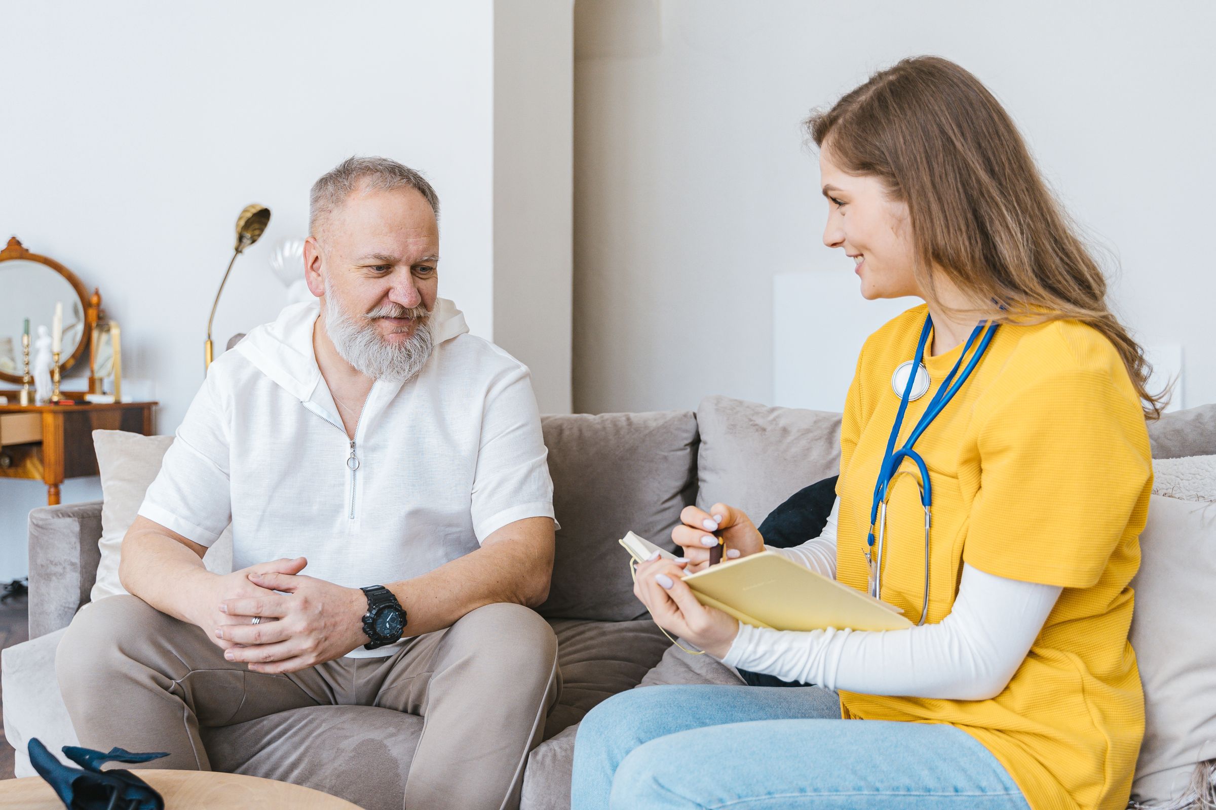A health worker takes notes while talking with an older man on his sofa at home