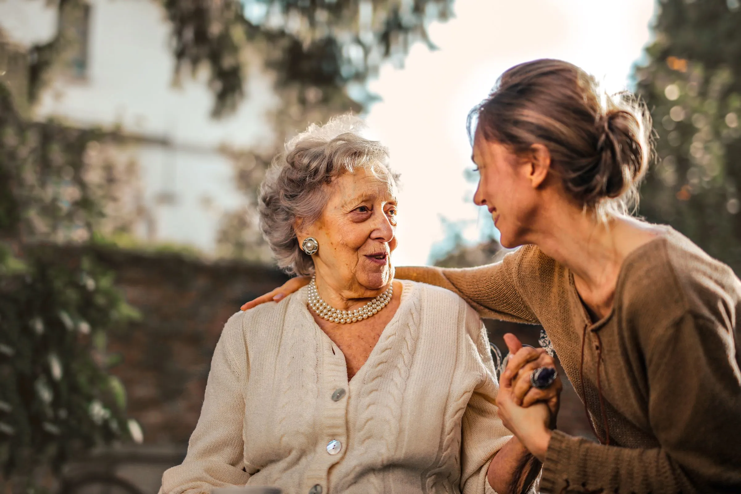 An older woman in a cream cardigan and pearls smiles as her adult daughter leans in, holding her hand, in a sunlit garden