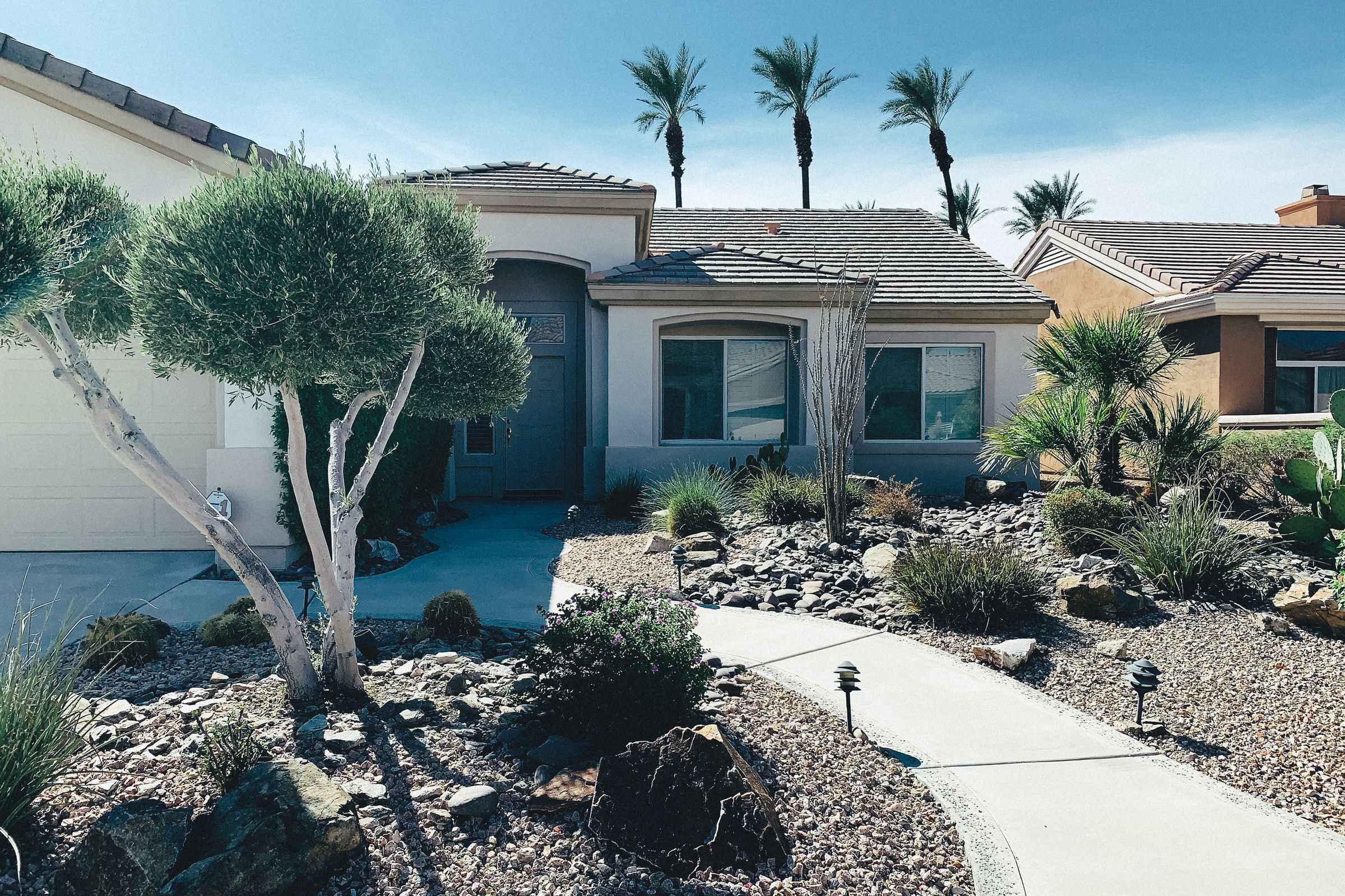 A single-storey stucco home with a tiled roof, palm trees and desert landscaping along the front walk