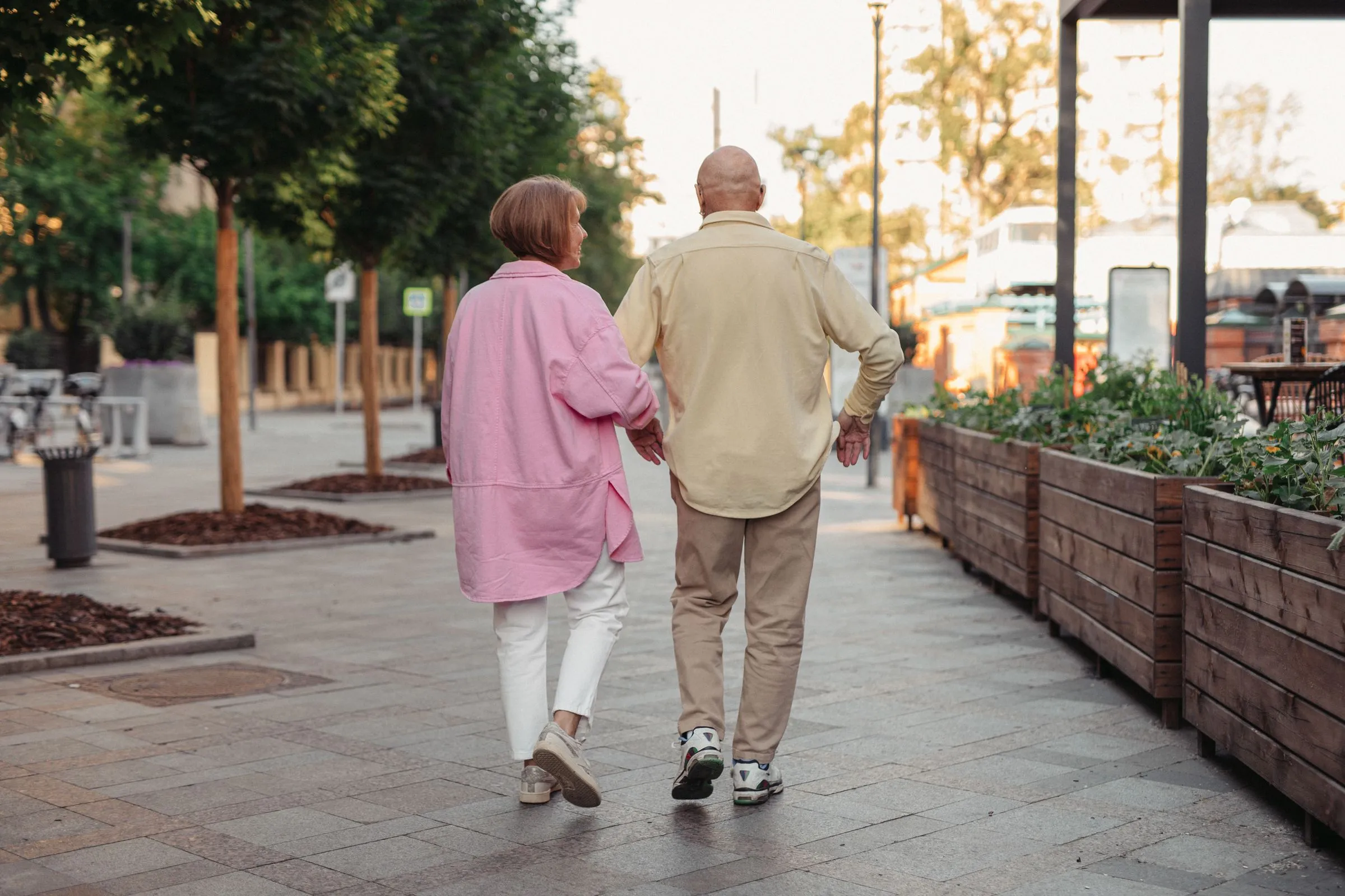 An older couple walk arm in arm along a tree-lined promenade on a sunny day