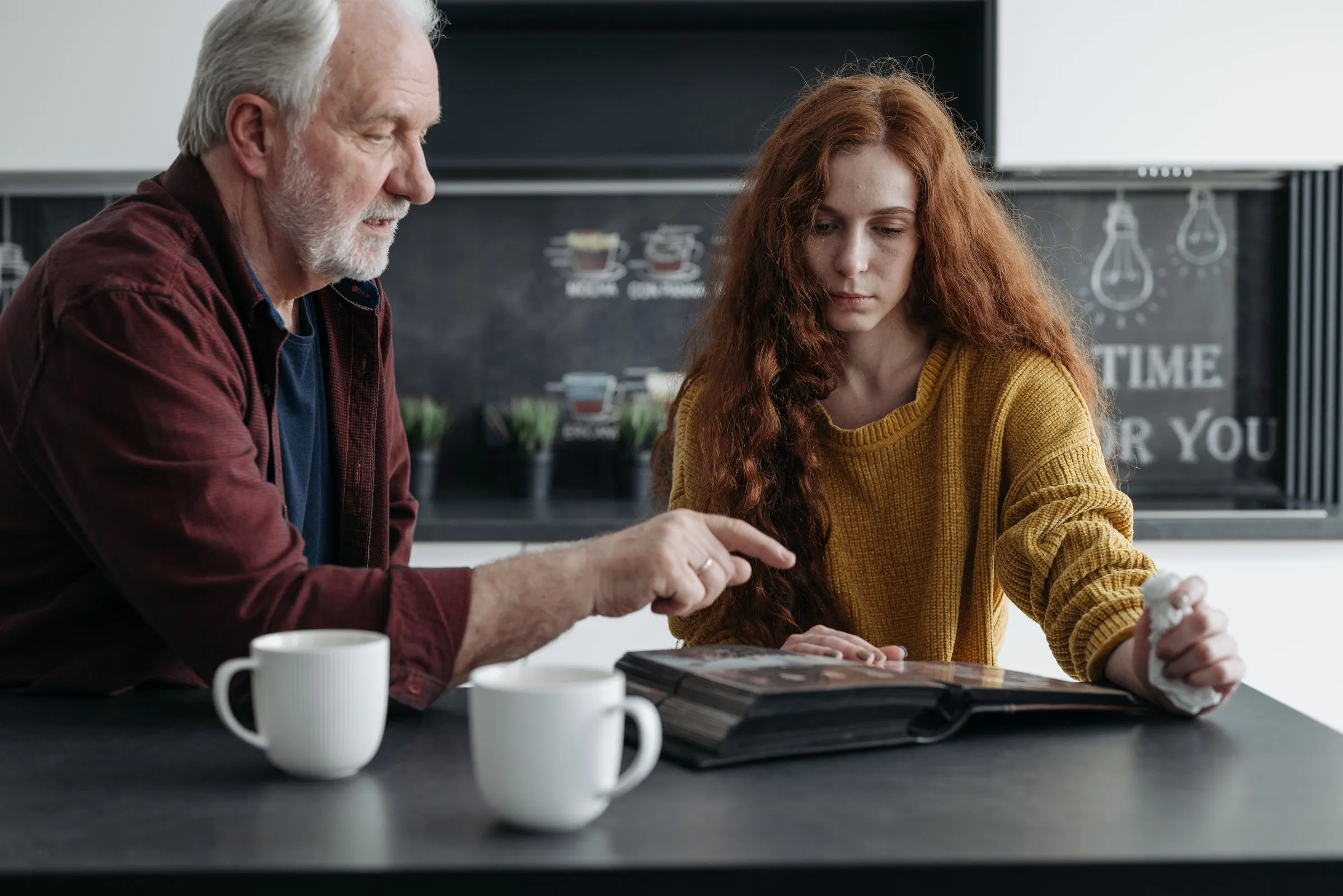 An older man points to a page in a photo album while his daughter looks on, coffee cups on the table