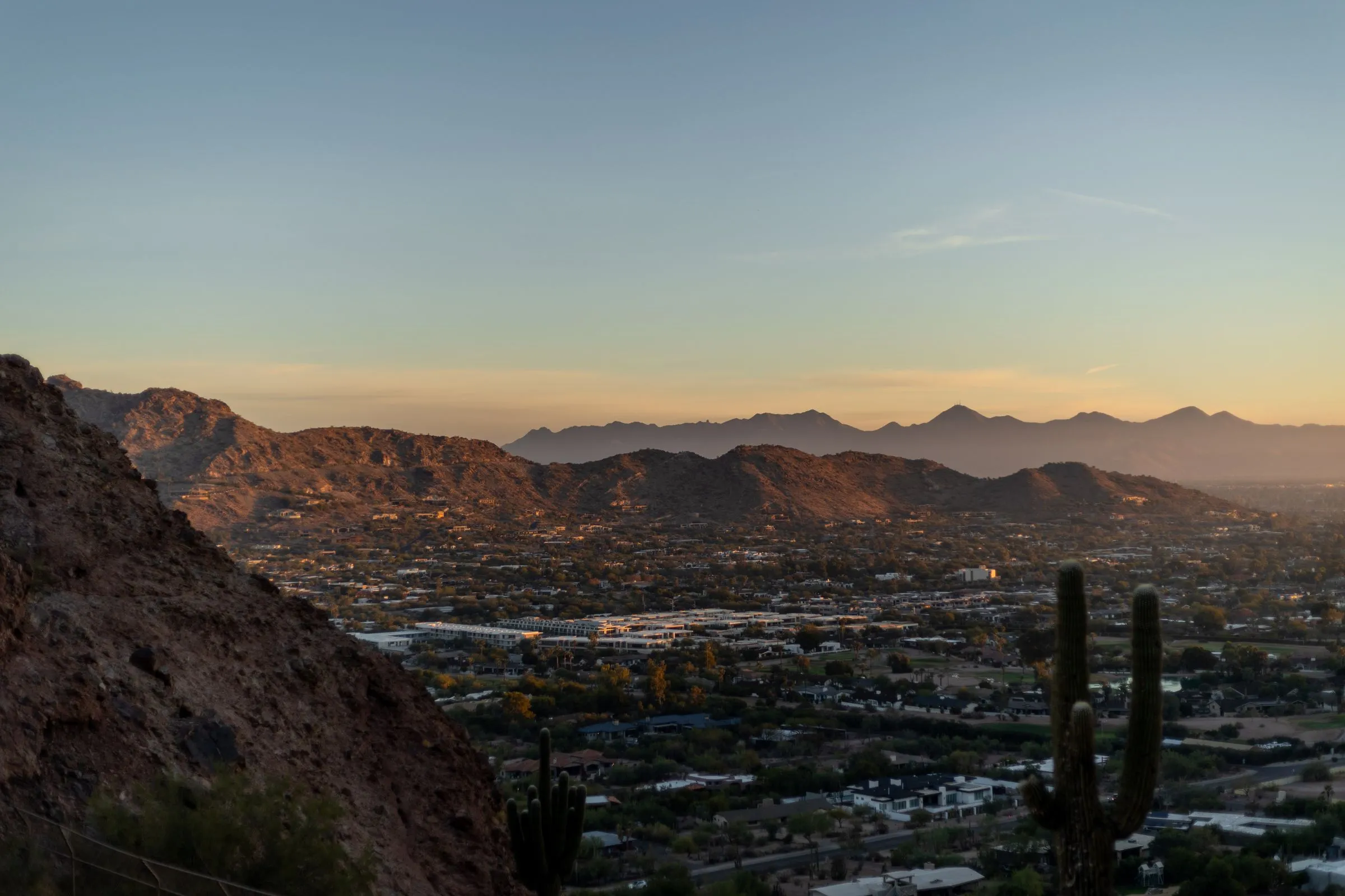 The Phoenix valley at sunset seen from a desert hillside, with a saguaro cactus in the foreground and mountains beyond