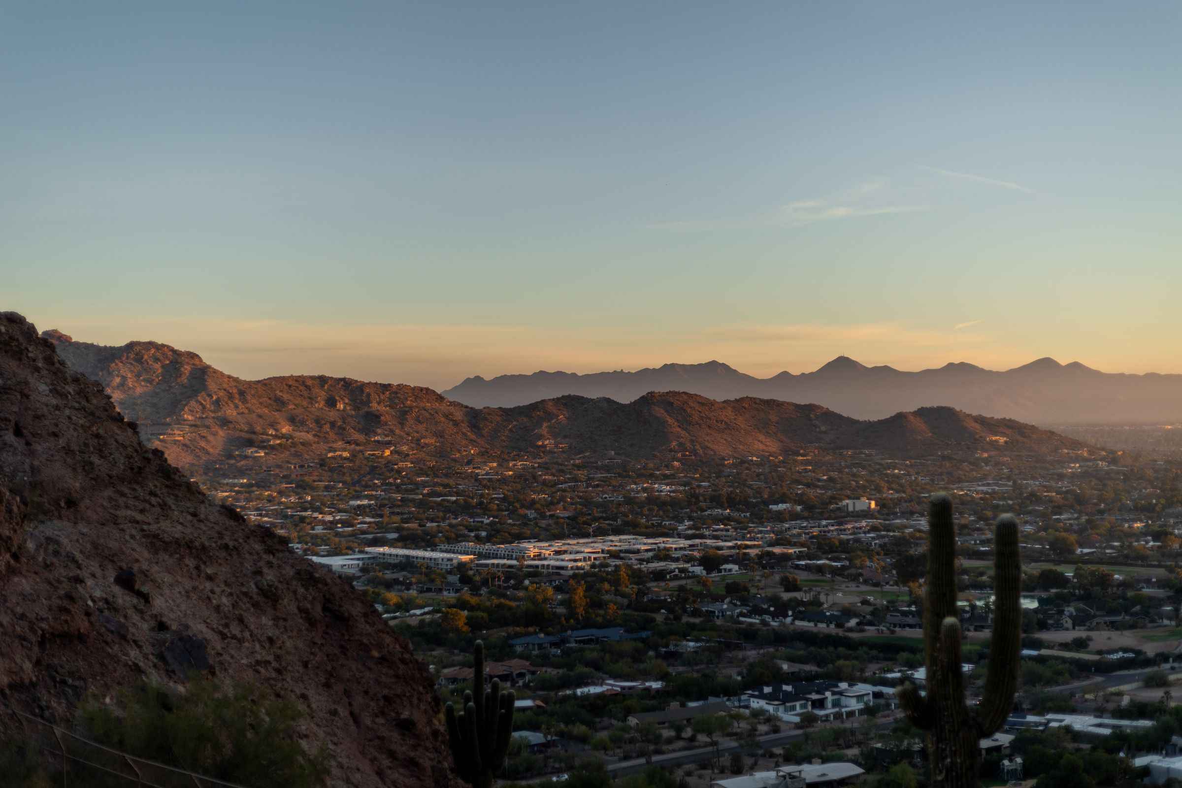 The Phoenix valley at sunset seen from a desert hillside, with a saguaro cactus in the foreground and mountains beyond