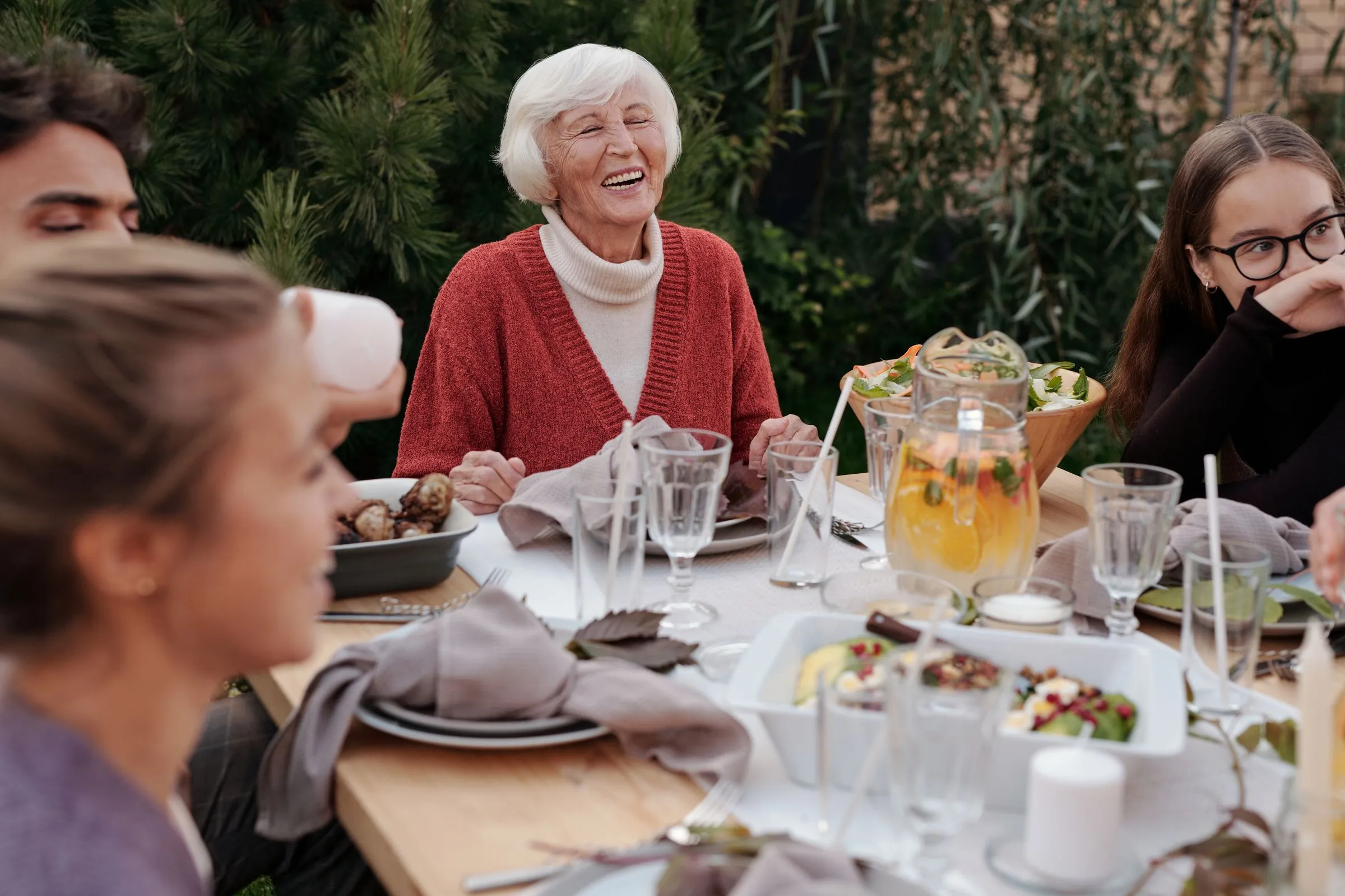 An older woman laughs with her family around a dinner table set outdoors in a garden
